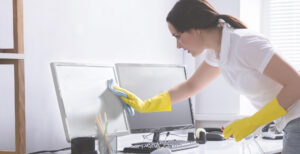 A woman wearing yellow gloves cleaning a computer monitor in an office using a cloth and spray bottle.