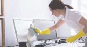 A woman wearing yellow gloves cleaning a computer monitor in an office using a cloth and spray bottle.